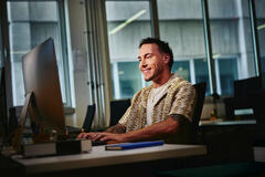 A young, smiling professional works at a computer in a modern office, illuminated by large windows in the background. He is wearing a distinctive patterned, short-sleeved collared shirt.