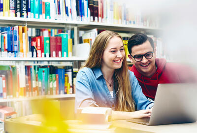 A woman with long, blonde hair and a denim shirt smiles while looking at a laptop. A man with short, dark hair, glasses, and a red hoodie is looking at the laptop over their shoulder, also smiling. They appear to be in a library, with bookshelves filled with books in the background.