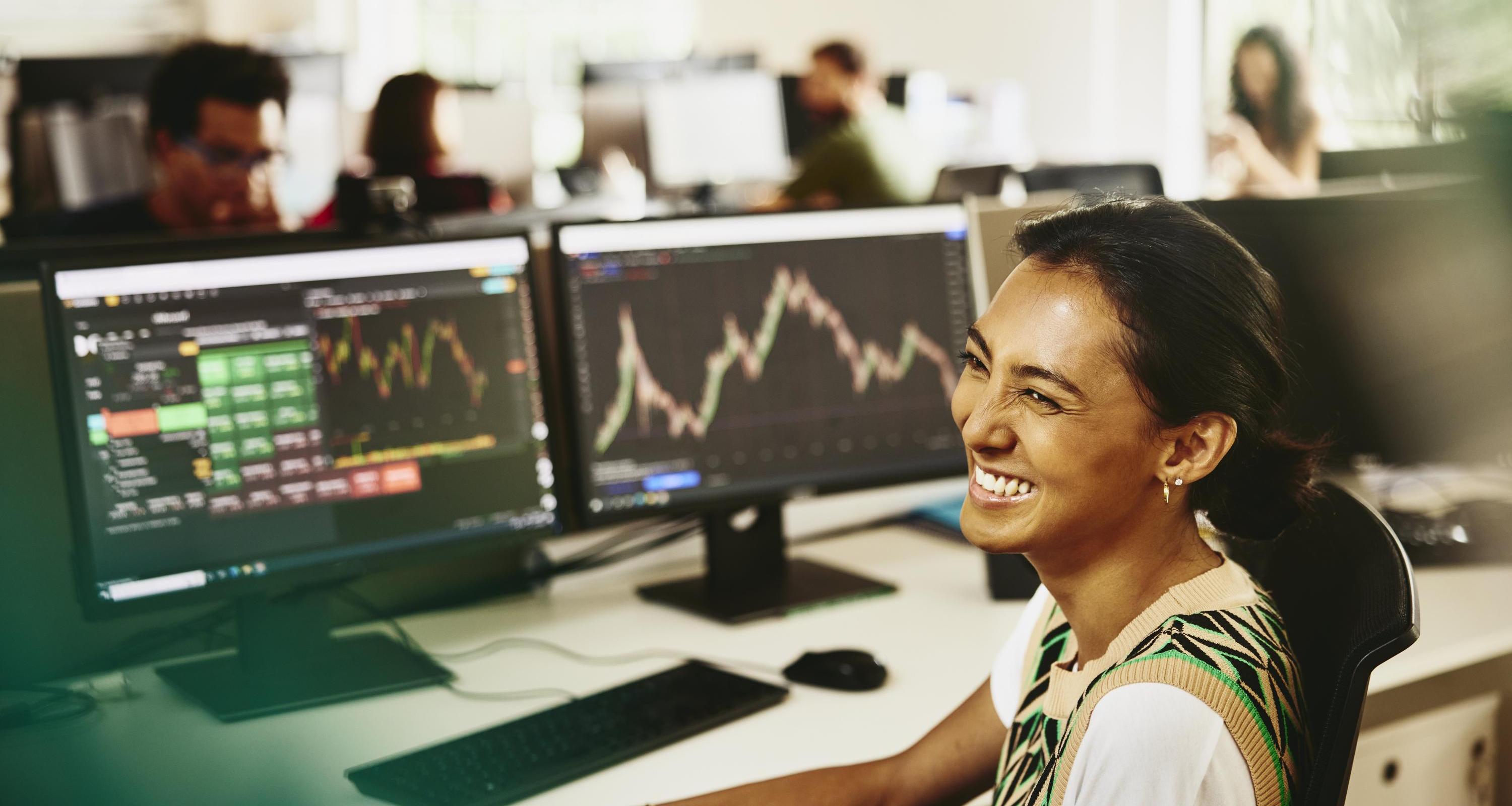 Woman smiling while sitting behind her desk, screens displaying financial information. Woman smiling while sitting behind her desk, screens displaying financial information.